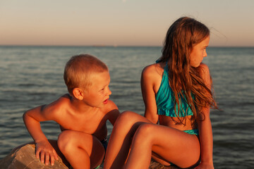 Brother and sister are sitting on the rocks on the seashore, at sunset. Girl with long hair, blond boy. Both looking at the sunset