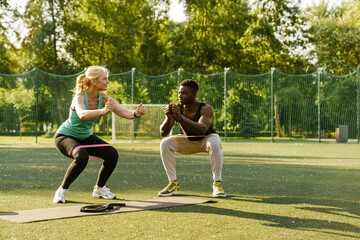 African trainer teaching mature woman doing squats with resistance bands outdoors in park