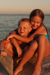 Fototapeta na wymiar Happy brother and sister are sitting on the rocks at the sunset. hugging and looking at the camera