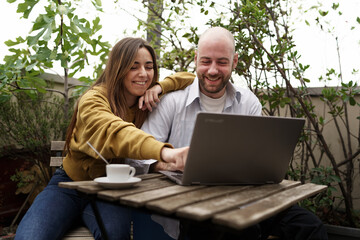 Young Couple Enjoying Coffee and Laptop at Outdoor Cafe - A bald man and long-haired woman laugh as...