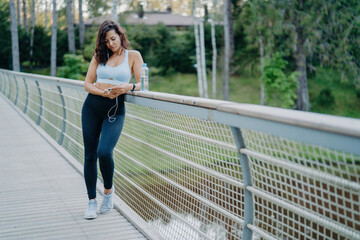 Slim European woman in active wear checks phone, listens to music with earphones on the bridge. Embracing the sport and lifestyle concept amid nature's backdrop.