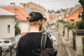 A man tourist in a cap, with a bun of hair and with a backpack explores the city looking at the panoramic view of the city, view from the back, copy space.