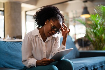 Beautiful African businesswoman sitting on sofa in office. Young tired woman using digital tablet.