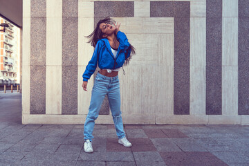 Photo shoot of stylish caucasian hip hop dancer posing having fun in blue jacket, jeans, black top and white tshirt, enjoy the urban town as background. City buildings shooting of moving fitness model