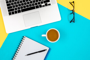 Notebook, laptop, eye glasses and cup of coffee on a blue and yellow background.