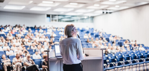 Female speaker giving a talk on corporate business conference. Unrecognizable people in audience at...