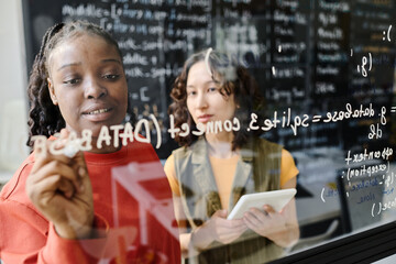 African American teacher writing codes on blackboard and explaining IT to student in the classroom