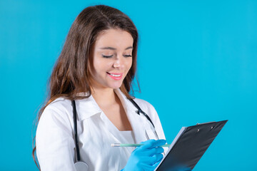 Young brunette doctor in a white coat with stethoscope making notes isolated on the blue background. 