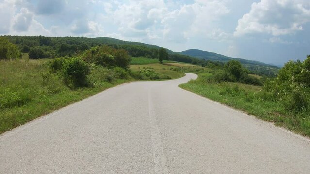 Driving a car or motorbike downhill on a winding country road, surrounded by corn fields and meadows. Beautiful sky with clouds, mountains in the distance. POV shot