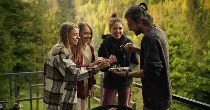 A group of friends try the food they cooked on the grill in a country house overlooking the mountains with a coniferous forest. The guy holds a plate of food and stands near the barbecue