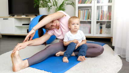 Fototapeta premium Cute baby boy sitting on fitness mat and looking at his mother stretching legs before sports training. Concept of healthcare, sports and yoga at home.
