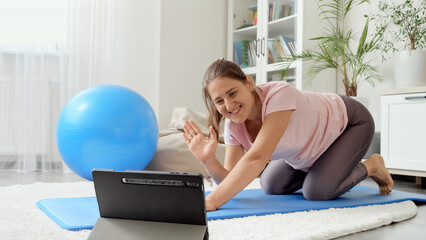 Happy smiling brunette woman having online fitness class waving in camera of tablet computer