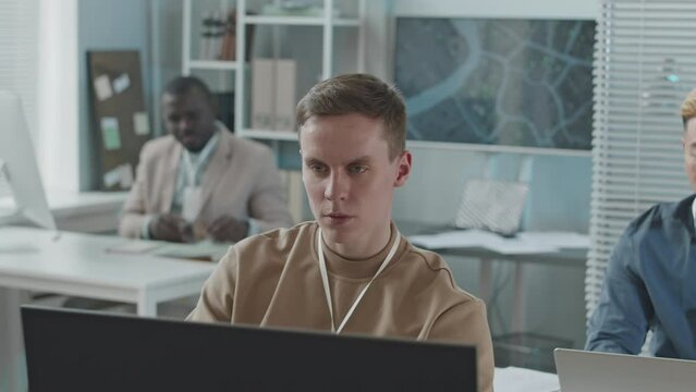 Chest up slowmo of young concentrated Caucasian male data engineer watching information on desktop computers while working in security agency together with diverse team of colleagues