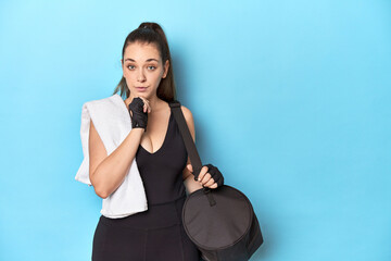 Young woman ready for gym with essentials on blue backdrop looking sideways with doubtful and...