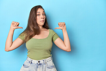 Young Caucasian woman in a green top on a blue backdrop feels proud and self confident, example to follow.