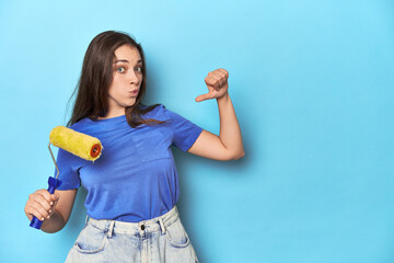 Young woman with yellow paint roller on a blue background feels proud and self confident, example...