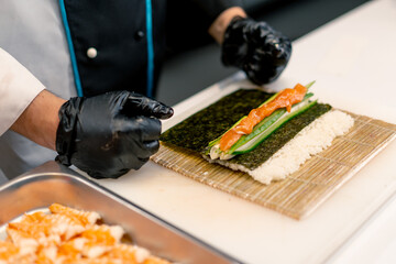 Close-up of a sushi maker in black gloves preparing sushi using rice nori sheets salmon fillet curd...