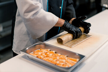 Close-up of a sushi maker in black gloves preparing sushi using rice nori sheets salmon fillet curd cheese cucumber avocado on the kitchen table 