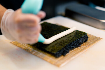 Close-up of a sushi chef spreading cream cheese on black rice on a nori sheet in the process of making rolls in the kitchen of a sushi restaurant