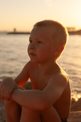 A blond boy on the seashore sits and looks thoughtfully to the side
