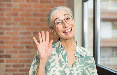 senior pretty woman smiling happily and cheerfully, waving hand, welcoming and greeting you, or...