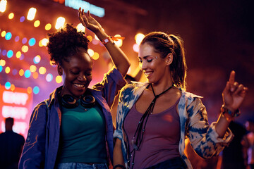 Happy female festival goers dancing in front of music stage at night.