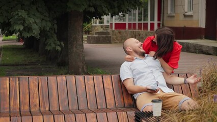 Man sitting on a bench and using smartphone, waiting for his girlfriend, playful woman in red dress make surprise, closing his eyes and kissing, lovers date in the summer city