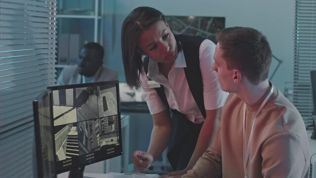 Medium slowmo of multiethnic couple of professional data engineers monitoring and discussing info on multiple outdoor CCTV surveillance cameras on computer desktop in dark office