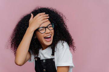 Ecstatic African American woman, curly hair, glasses, laughs sincerely, isolated on purple wall.