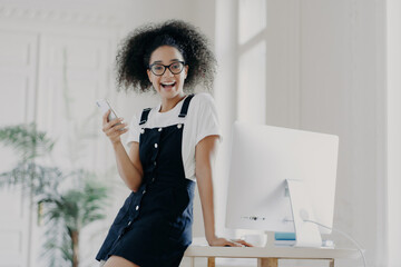 Joyful female financier, mobile in hand, salary received on time, by wooden table, working on computer, wearing white t-shirt and sarafan, happily texting.