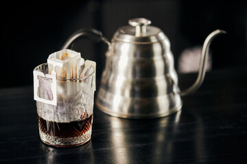 glass with paper filter bag and black expresso near blurred drip kettle on black table, drip method