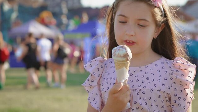 Happy joyful cute little girl eating ice cream at amusement park on defocused people and carousels background. Having fun and relaxing at weekends in luna park outdoor
