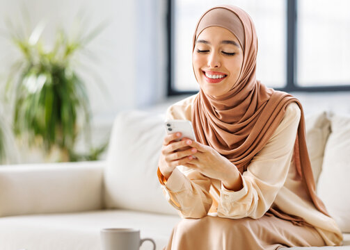 Beautiful overjoyed young smiling muslim woman in traditional religious hijab  reading great news on smartphone.