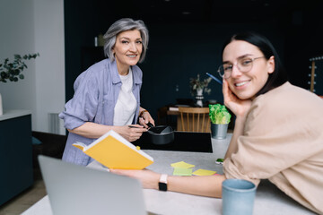 Cheerful mature woman standing with coffee pan at kitchen while daughter joining with recipe