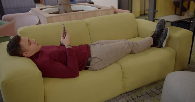 Young male worker resting in the cozy couch inside the office lounge area, trucking
