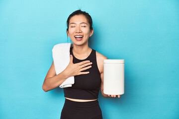 Young Asian sportswoman with protein bottle, gym setup, laughs out loudly keeping hand on chest.