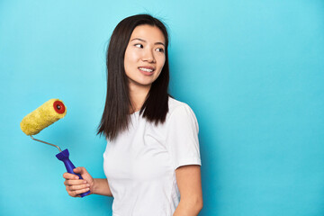 Young Asian woman with paint roller, DIY concept, looks aside smiling, cheerful and pleasant.