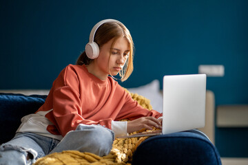 Focused teen girl wearing wireless headphones sitting on sofa with laptop, studying online, having virtual lesson. Child teenager surfing internet, learning something new 