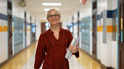 Closeup portrait of pretty mature woman teacher holding books walking down an empty school hallway.