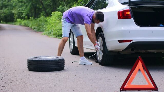 A young man sets the spare wheel of his white car on the side of the road or the edge of the road. Replacing a wheel on a car with an emergency sign on the road.