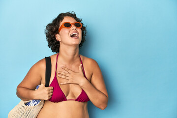Young woman in bikini with beach bag laughs out loudly keeping hand on chest.
