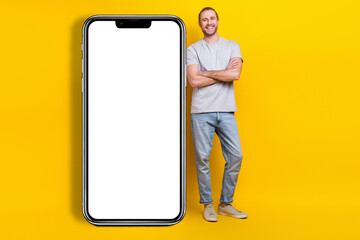 Full size photo of satisfied man gray t-shirt denim pants standing near white ui panel arms crossed isolated on yellow color background