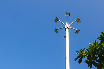 Low-angle view of lights pole in the middle of the park with green trees foreground and blue sky background. Lights pole isolated on blue sky.