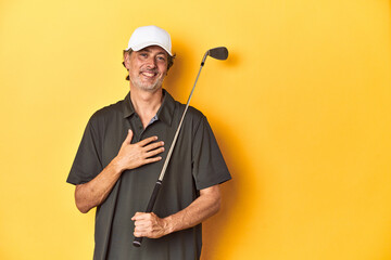 Middle-aged golfer with club and cap on yellow laughs out loudly keeping hand on chest.