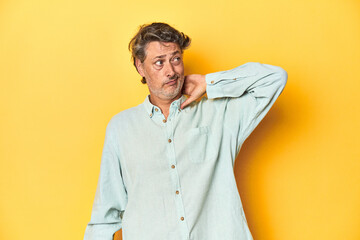 Middle-aged man posing on a yellow backdrop touching back of head, thinking and making a choice.