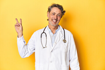 Middle-aged doctor standing against a yellow backdrop joyful and carefree showing a peace symbol with fingers.