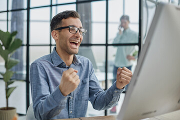 Excited modern businessman sitting at office desk and rejoicing his success