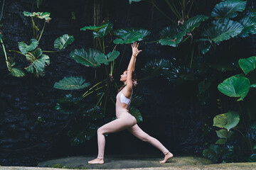 Side view of young woman feeling inspiration during hatha workout training at beautiful nature environment with greenery leaves, Caucasian fit girl in sportswear meditate in asana pose during yoga