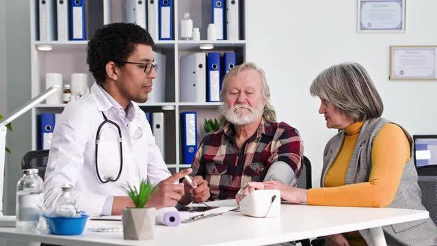 reception of patient, black doctor measures pressure of an old man, a loving elderly wife supports husband in hospital office