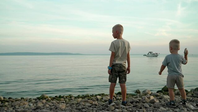 Two little sibling brothers kids children on vacation at sea or ocean. Two Caucasian children at soft sunset or sunrise stand on rocky shore of sea or ocean.